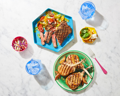 Overhead shot of grilled porkchops on a green plate and grilled pork loin on a blue plate sitting on a white marble table top.