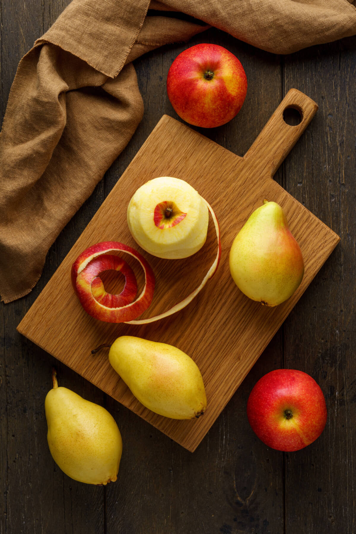 Apples and pears on a cutting board with one apple that has been almost fully peeled, with the peel beside it